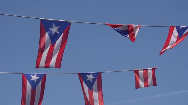 A string of Puerto Rican flags waving in the wind against a clear blue sky with sun rays.
