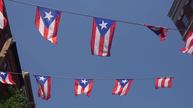 A string of Puerto Rican flags waving in the wind against a clear blue sky with sun rays.
