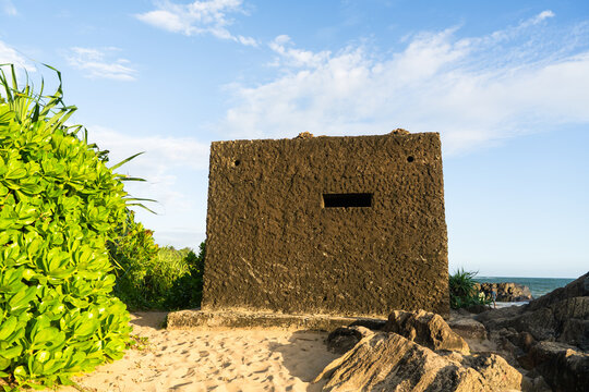 View of a historic concrete bunker on a sandy beach surrounded by lush green vegetation and coastal rocks under a clear blue sky in Galle, Southern Province, Sri Lanka.