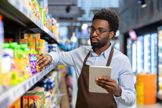 Male grocery worker in an apron organizing products on a supermarket shelf while accurately monitoring stock levels and managing inventory using a modern digital tablet