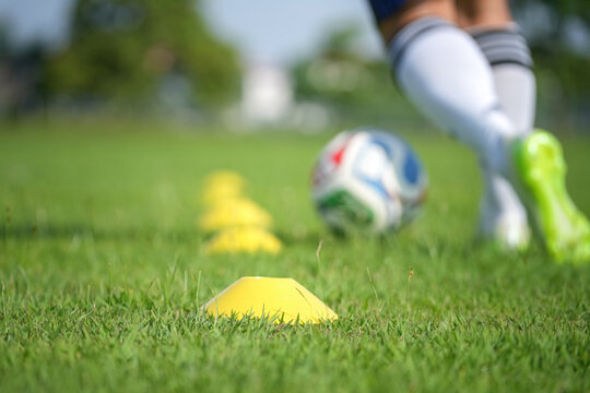 A football player is training drillbing the ball around the speed cone on grass pitch, close-up at the speed cone on foreground and player as blurred backgound.