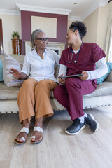 Fototapeta premium Senior African American woman sitting at home chatting with care worker in scrubs holding tablet