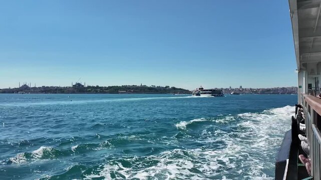 Scenic view of Istanbul Bosphorus from a ferry on a sunny day, with waves, passing boats, and the historical peninsula skyline featuring Hagia Sophia and Topkapi Palace