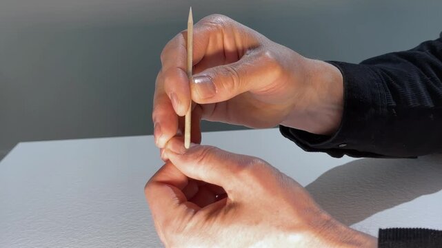 Hands hold a wooden cuticle stick over a fingernail in close up. The fingers adjust the tool during nail care on a plain surface. Salon manicure treatment and grooming concept..