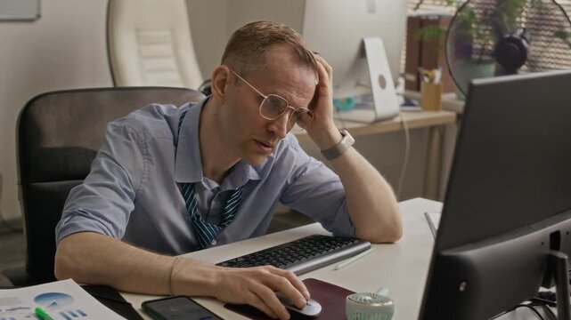 Medium shot of overheated man sitting at desk, taking off glasses and wiping sweat while trying to work in stuffy office