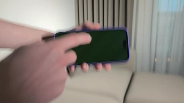 Hand holds smartphone with green screen in a living room. The device stays horizontal in close up with slight motion across the clip. Mockup for app design, advertising, and video editing..