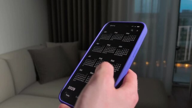 Hand holds smartphone with calendar app in close up. The vertical mobile screen scrolls through yearly dates in a living room. Planning, scheduling, and digital time management concept..