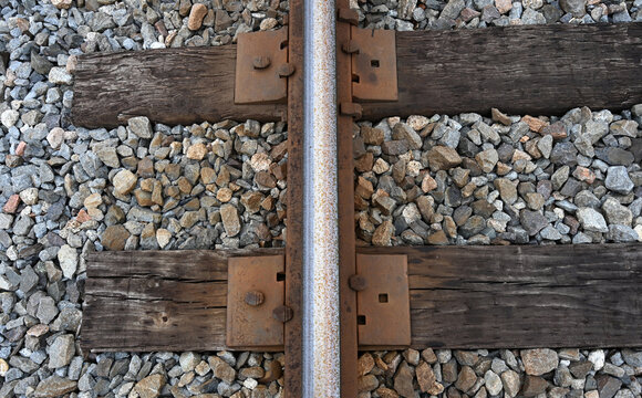Horizontal close up of a weathered railroad rail with rusty tie plates, rail spikes, two cracked wood ties, rust pits, mixed ballast gravel, wood chips, and dried leaves