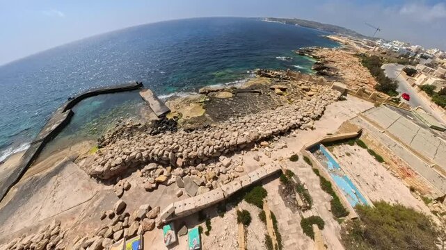 Drone flying backwards over the abandoned and ruined resort hotel in Marsaskala in Malta with San Tumas watchtower in the background.