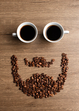 Creative top view of a face with beard and mustache made of coffee beans and two coffee cups as eyes on a wooden table