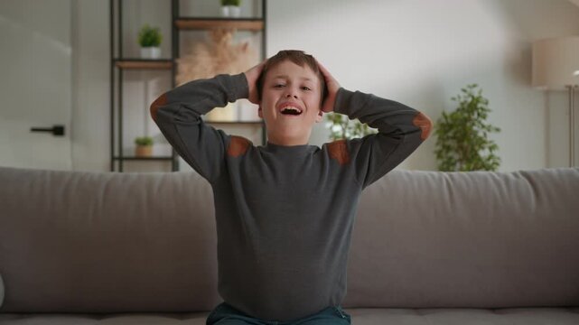 Young boy expresses extreme frustration and distress by screaming with hands on head while sitting on sofa. The intense moment highlights childhood emotions, agitation and the raw energy of outburst