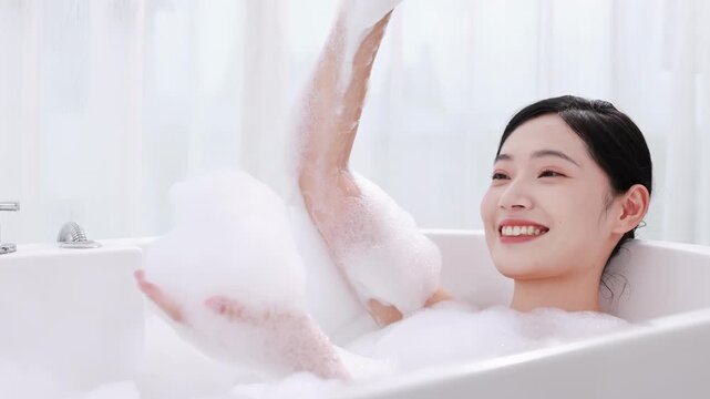 Attractive elegant seductive woman blowing off white foam while washing in luxury bathroom, girl enjoying luxury bath romantic happy girl taking bathtub plays blowing on bubbles, sensual water care