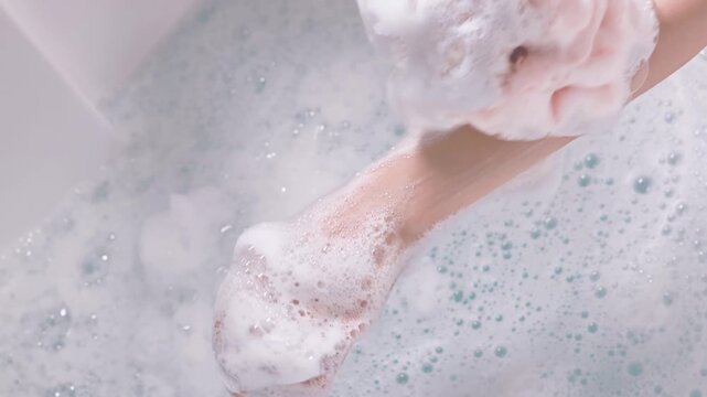 Unrecognizable young woman washes her arm with sponge and body wash suds while relaxing in hot bath with bubbles in a bathtub filled with foam inside a hotel room