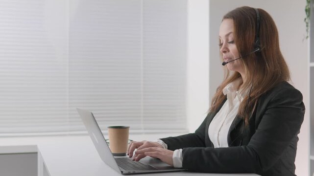 Focused female support agent communicates with clients via headset while working on her laptop in a bright office. She demonstrates remote work efficiency and digital communication skills