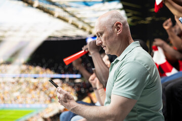 Fototapeta premium Focused football fan placing live bet ticket through mobile gambling app during soccer match with cheering supporters at the stadium. Man experiencing high risk betting tension with phone.