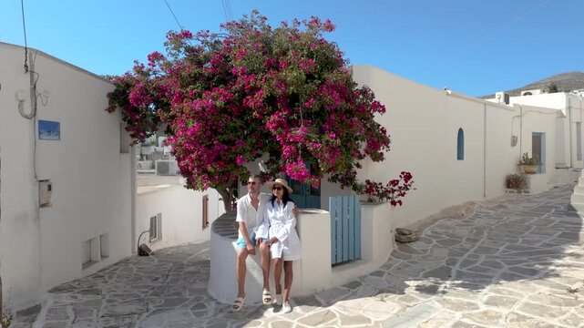 A diverse couple discovers the enchanting streets of Paros, Greece as a traveler enjoys a moment of relaxation beneath a blooming bougainvillea. A perfect blend of culture and natural beauty awaits.