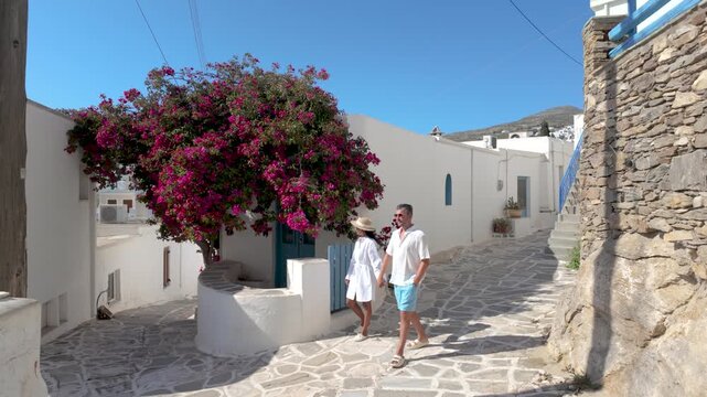A diverse couple wanders through the picturesque streets of Paros, Greece, where vibrant bougainvillea blooms adorn whitewashed walls. Relax and soak in the sunny atmosphere of the Cyclades