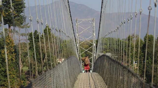 Surkhet, Nepal - 06 January 2024: View of a person carrying a basket while walking across a long suspension bridge surrounded by greenery and distant mountains.