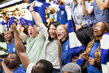 Fototapeta premium Passionate blue women supporters cheering with raised arms in packed stadium stands during intense soccer match atmosphere. Sports fans celebrate every goal, football culture equality.