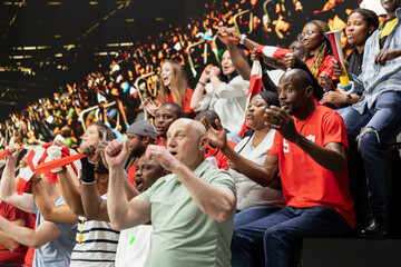 Fototapeta premium Ecstatic football supporters cheering and waving scarves after dramatic goal during soccer match in crowded stadium stands. Joyful crowd reaction, raised fists and jumping fans.