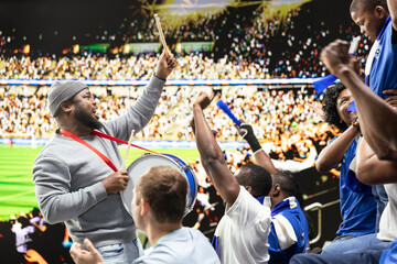 Fototapeta premium Passionate football capo beating drum and directing blue supporter group from stadium stands during coordinated chant at live soccer match. Hardcore fans wave scarves, stadium chants.
