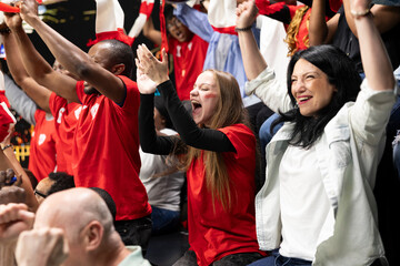 Fototapeta premium Female soccer fans celebrating goal with raised arms in crowded stadium stands, surrounded by red team supporters and energetic live match atmosphere in fan section. Pure joy and excitement.