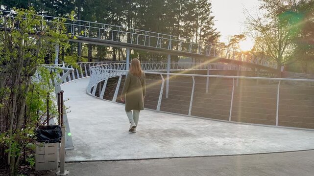 Young woman walking on a modern curved pedestrian bridge in a public park at sunset