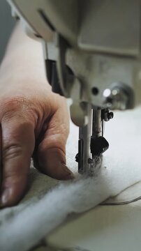 Vertical video. Industrial sewing machine stitching soft fabric in textile workshop. Close-up shot of moving needle, fabric edge, sewing process and hands guiding material. Filmed in 4K slow motion.