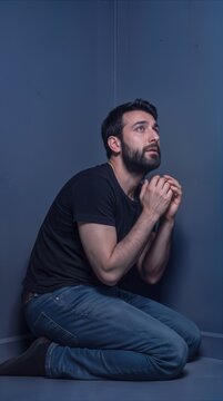 Pleading man kneeling in corner of dark room. Spiritual prayer or desperate request for help.