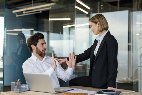 Businessman reacting uncomfortably, raising hands to stop a confident female coworker sitting on his desk, creating an awkward situation in a corporate workplace