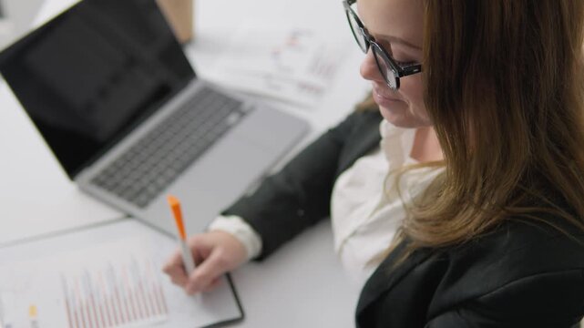 Confident female professional working on financial analysis, using calculator, reviewing business documents. Office workspace with laptop, clipboard, financial charts for effective decision making