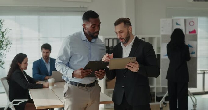 Portrait of African American and Middle Eastern businessmen check tablet in office as team works near desk. Colleagues compare data and discuss project