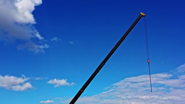 Construction crane boom with a hook extending into a vivid blue sky with fluffy white clouds