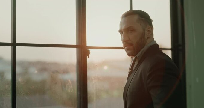 Portrait of Middle Eastern businessman near window turns toward camera. Bearded executive looks over shoulder and pauses between meetings while considers plans