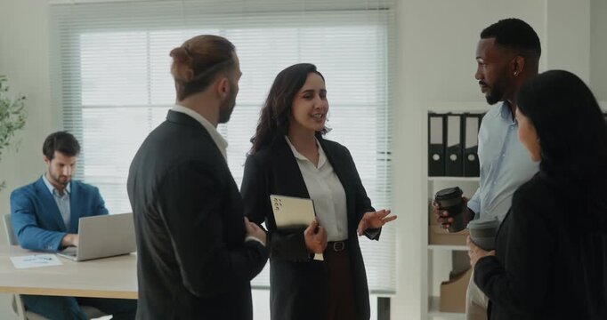 Portrait of young Latin woman boss talks to diverse professionals team in workspace. Hispanic lady manager holds tablet and informs colleagues in office