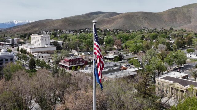Nevada and American flag hanging on tall flagpole overlooking historic brick neighborhood in Carson City, Nevada with green trees andSierra Nevada mountains under clear sky in USA. Aerial wide shot.