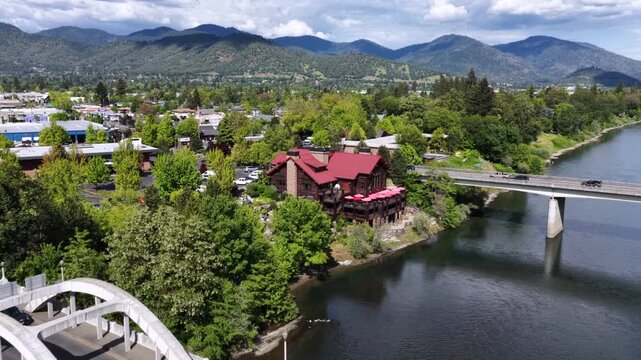 Grants Pass Oregon white arch bridge with active car traffic crossing blue Rogue River, red-roof historic building among lush green trees, second bridge visible in background. Aerial flyover shot,