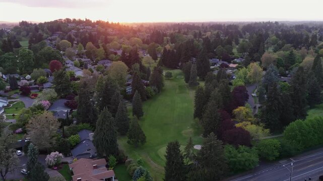 Suburban neighborhood with golf course fairway, dense spring trees, single-family homes, and warm golden sunset light glowing along horizon. Eugene, Oregon, USA. Aerial wide shot.