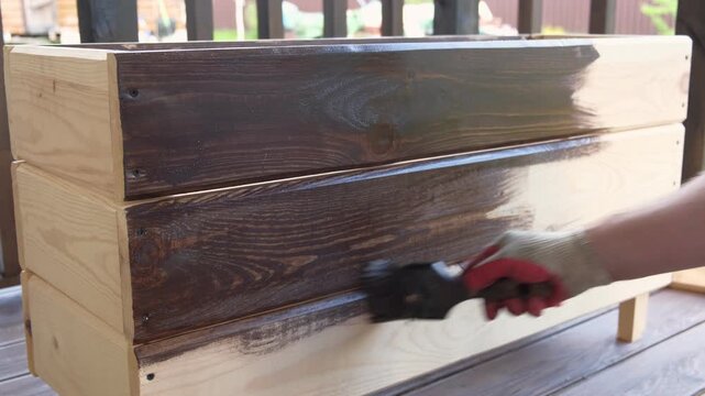 Brown oil for protection of wood by staining. Painting fence for protecting and pests. Woodworker applying finish to pine board box. Repairman varnishing with brush wooden fencing wall plank surface.