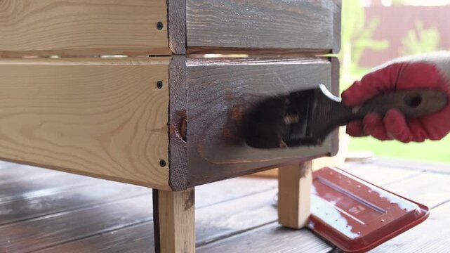 Brown oil for protection of wood by staining. Painting fence for protecting and pests. Woodworker applying finish to pine board box. Repairman varnishing with brush wooden fencing wall plank surface.
