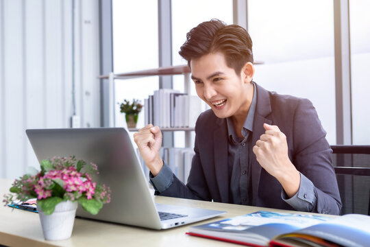 Happy of asian young businessman see a successful business plan on the laptop computer and pen on wooden table background in office,business expressed confidence embolden