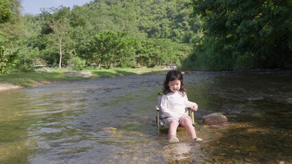 Fototapeta premium A young child sits peacefully on a camping chair in a shallow river, enjoying a quiet moment of sensory play and connecting with the natural environment.