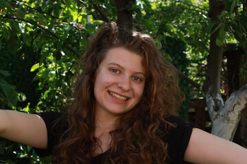 Fototapeta premium Young woman with curly hair smiles while standing in a garden during a sunny day surrounded by green leaves and trees