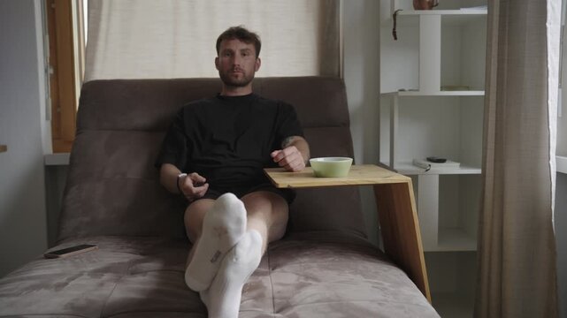 Young bored man watching tv, changing channels with a remote, and eating from a bowl at home