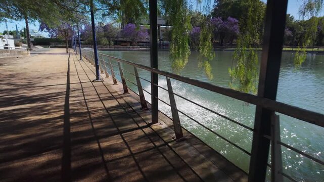 Sunny Day on a Picturesque Riverwalk in Sevilla with a Boat Creating a Serene Wake Along the Railing