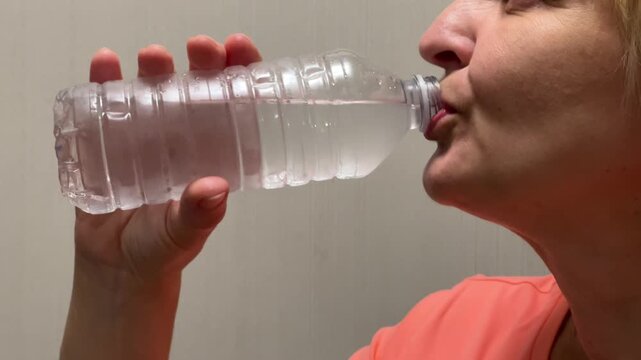 A person sips water from a plastic bottle in a plain room. Shows the act of hydration during a regular day. Clear focus on drinking.