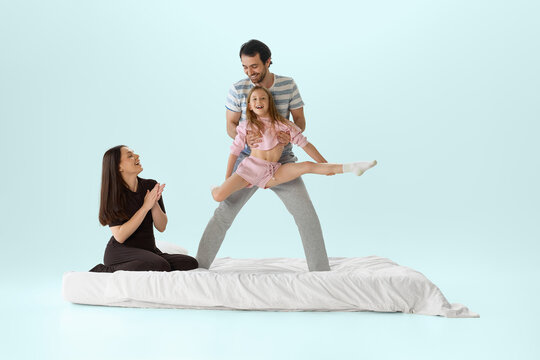 Father holding daughter in split jump above white bed while mother applauds. Concept of childhood confidence, family support, active play, bedroom lifestyle, and mattress comfort.