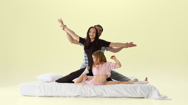 Playful family posing like dancers on white bed in pastel yellow studio. Concept of family creativity, active childhood, parenting fun, bedroom lifestyle, and joyful bonding.