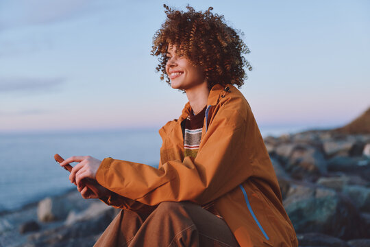 Cheerful woman lifestyle outdoors, smiling with curly hair, wearing a brown jacket, sitting on rocky coast, gentle sea breeze, sunset light, soft pastel sky relaxed pose and laughing