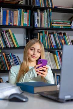 Teenage girl using smartphone while studying with laptop and books in library. Digital distraction, procrastination, lack of focus and modern education challenges concept.